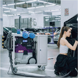 A woman technician kneels beside a car in a service garage while using a Sandwox tool cart with dust extractor and tool holders. The garage has bright overhead lights, other vehicles, and a no-smoking sign in the background.